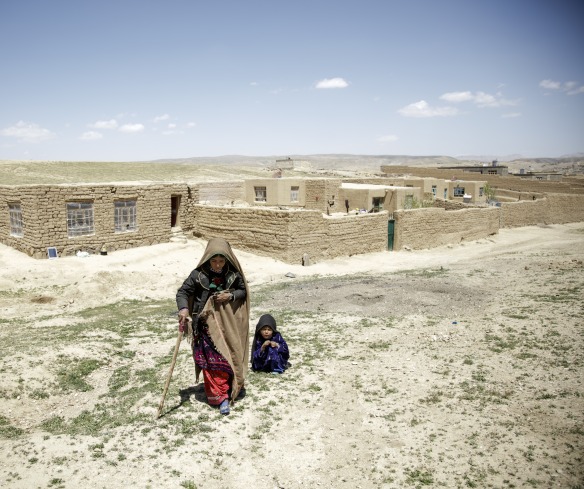 A mother walks away from her home with the help of a cane. Her daughter crouches behind her.